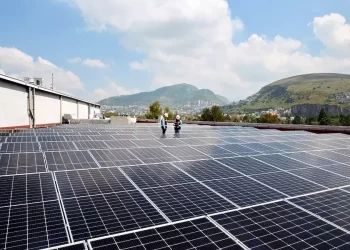 Rooftop solar panels stretch across a flat roof while two workers in safety gear inspect them; hills in background.