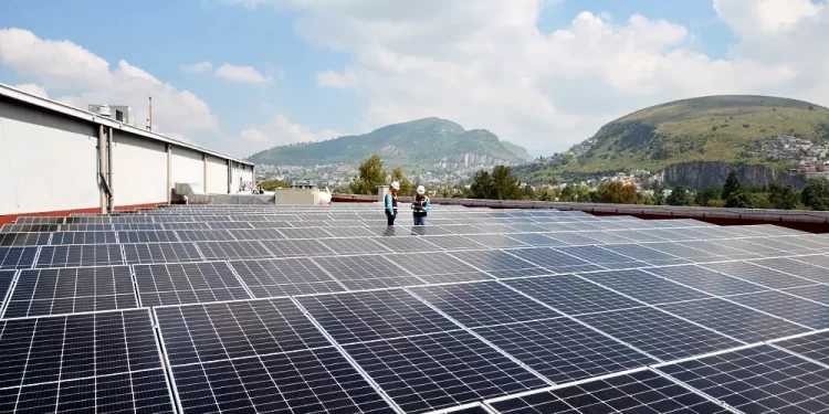 Rooftop solar panels stretch across a flat roof while two workers in safety gear inspect them; hills in background.