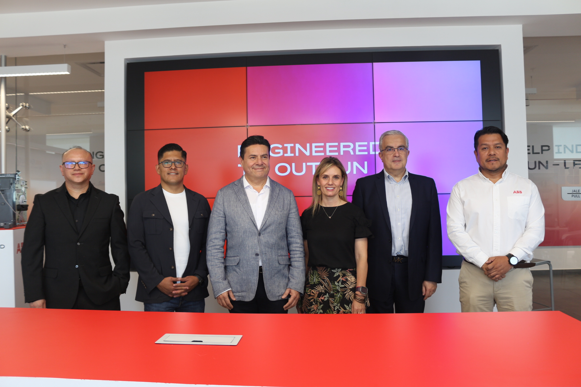Six professionals stand side by side behind a bright red table in a modern office, posing for a group photo in front of a colorful multi-panel display.