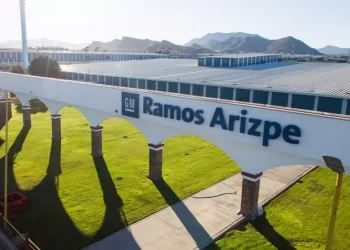 Sign on a white wall reading 'GM Ramos Arizpe' with arched colonnades and a green lawn in the foreground, factory buildings in the background and desert mountains beyond.