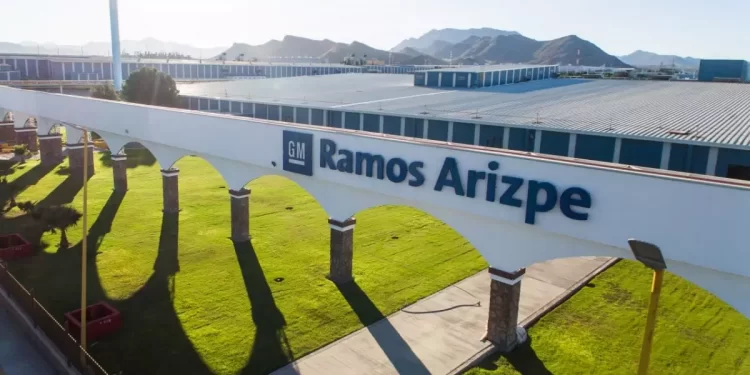 Sign on a white wall reading 'GM Ramos Arizpe' with arched colonnades and a green lawn in the foreground, factory buildings in the background and desert mountains beyond.