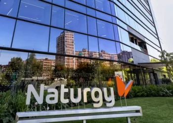 Front of a modern glass office building with a large Naturgy sign and orange logo in a landscaped area on a sunny day; reflections in the glass panels nearby.