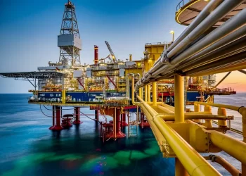 Offshore oil rig with yellow piping and platform extending over calm sea at sunset, illuminated by industrial lights