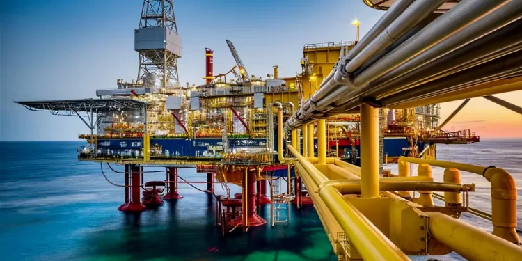 Offshore oil rig with yellow piping and platform extending over calm sea at sunset, illuminated by industrial lights
