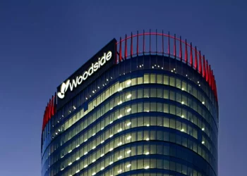 Cylindrical glass office tower at dusk with illuminated windows and a Woodside logo at the top-left.