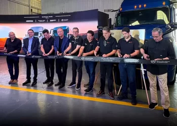 Nine men in dark shirts cut a ribbon at a truck company ceremony beside a blue semi-truck in a warehouse setting.