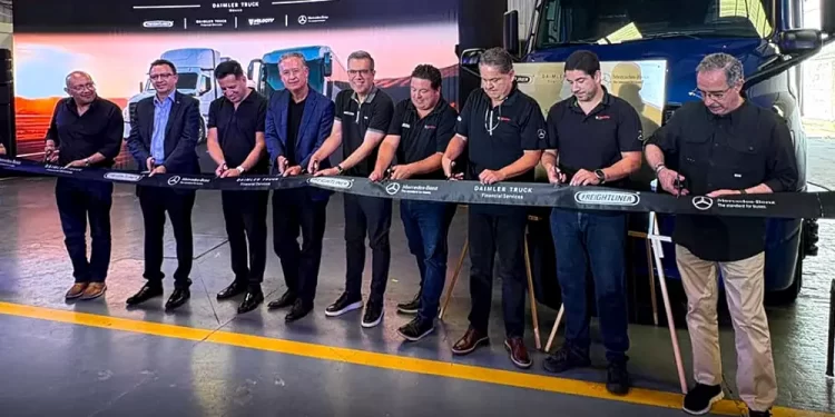 Nine men in dark shirts cut a ribbon at a truck company ceremony beside a blue semi-truck in a warehouse setting.