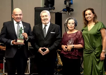 Four adults in formal attire stand together on a stage; the man on the left holds a small trophy/statue.