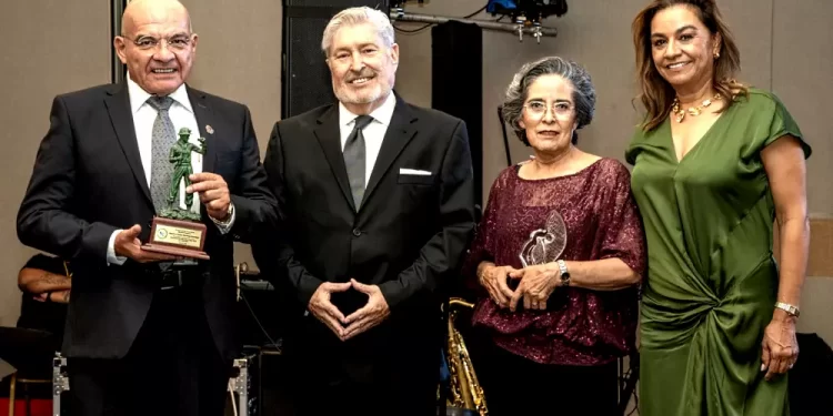 Four adults in formal attire stand together on a stage; the man on the left holds a small trophy/statue.