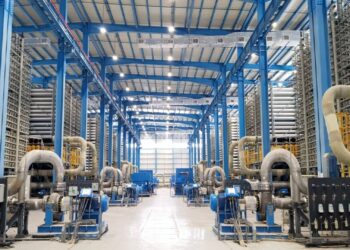 Industrial facility with blue steel framework and rows of large gray pipes and machinery along both sides of a spacious floor.