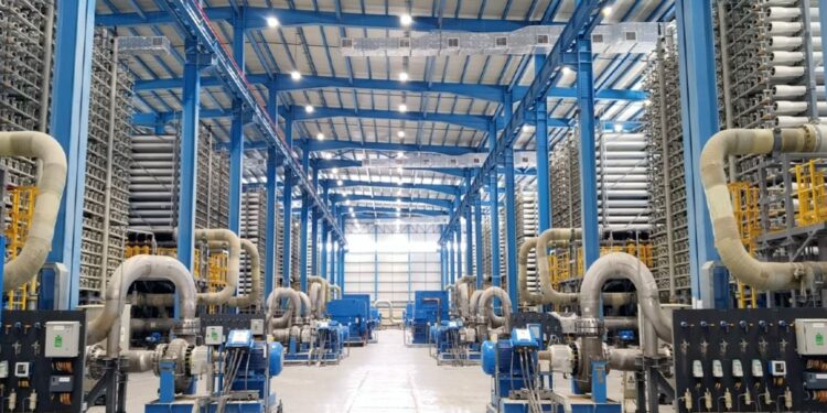 Industrial facility with blue steel framework and rows of large gray pipes and machinery along both sides of a spacious floor.