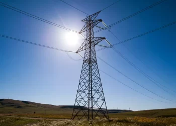 High-voltage transmission tower with power lines stretching across a sunny blue sky over a rural hill landscape.