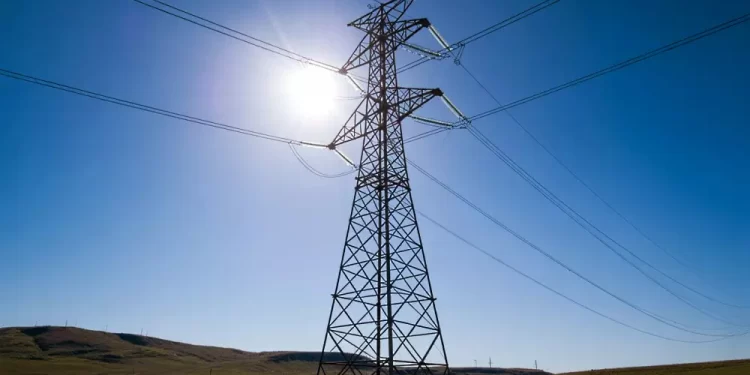 High-voltage transmission tower with power lines stretching across a sunny blue sky over a rural hill landscape.