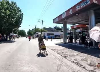 A man pushing a shopping cart with a yellow box along a sunny street beside a 24-hour gas station.','Street scene with a man pushing a cart, motorcycles in the road, and a 24-hour gas station on the right under a red canopy.','People along a sunny urban street; a man with a cart walks toward the camera near a gas station.','A busy small-town street scene showing pedestrians, a motorcycle, and a prominent 24 Horas gas station canopy.','Elderly man with a cart in the road while a gas station and parked bikes are visible on the right.