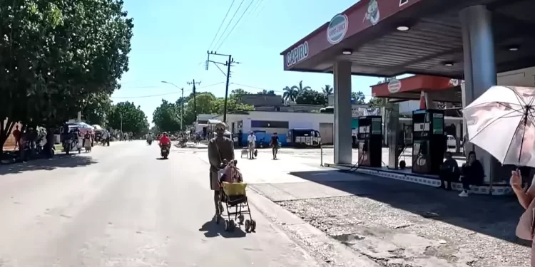 A man pushing a shopping cart with a yellow box along a sunny street beside a 24-hour gas station.','Street scene with a man pushing a cart, motorcycles in the road, and a 24-hour gas station on the right under a red canopy.','People along a sunny urban street; a man with a cart walks toward the camera near a gas station.','A busy small-town street scene showing pedestrians, a motorcycle, and a prominent 24 Horas gas station canopy.','Elderly man with a cart in the road while a gas station and parked bikes are visible on the right.