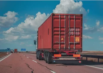 Red shipping container on the back of a semi-trailer driving on a highway under a blue sky and clouds. 110 characters? approximately fine to be concise.