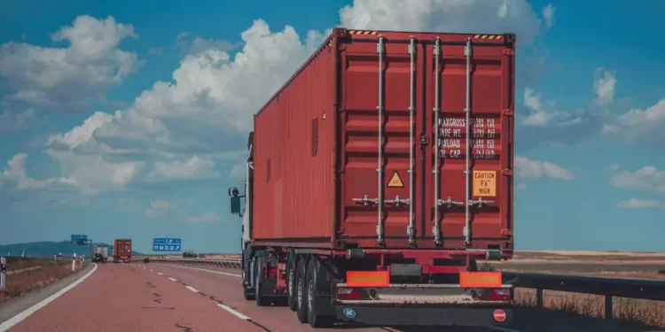 Red shipping container on the back of a semi-trailer driving on a highway under a blue sky and clouds. 110 characters? approximately fine to be concise.